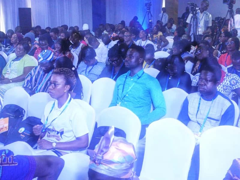 A large audience of professionals, likely health workers, seated in a conference hall during a formal event. Many attendees are wearing lanyards and some are looking toward a stage or speaker, with media cameras and tripods visible in the background.