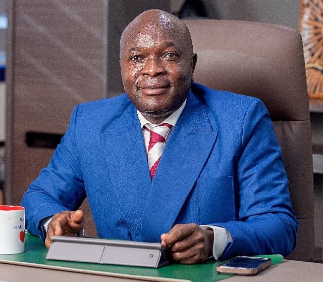 A portrait of Alhaji Seidu Agongo, the founder of Class Media Group, seated at an office desk. He is wearing a vibrant blue suit with a red and white striped tie and looking directly at the camera with a subtle smile.