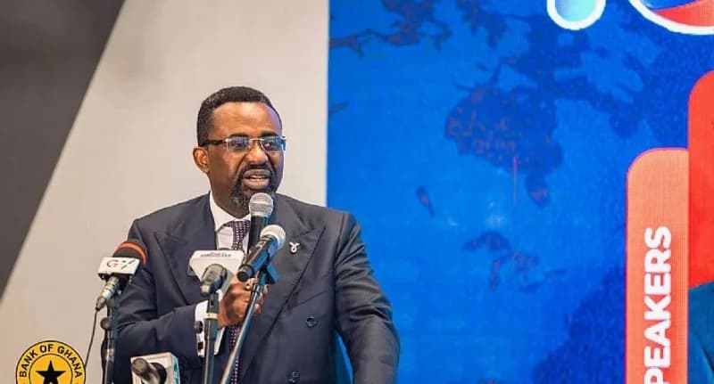 Dr. Ernest Addison, Governor of the Bank of Ghana, is pictured speaking at a formal event behind a podium with several microphones. He is dressed in a dark double-breasted suit and glasses, standing against a backdrop of a blue world map and a red 'SPEAKERS' banner. The Bank of Ghana logo is visible in the bottom left corner, indicating the official nature of the address.