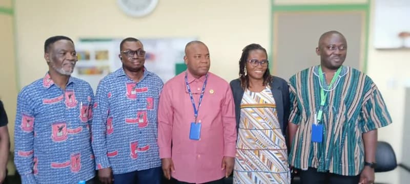 A group of five individuals, representing Wallahs Academy SHS and Ho Teaching Hospital, pose for a photo indoors. They are dressed in a variety of attire including school-branded patterned shirts, a pink formal shirt, and a traditional green-striped smock (fugu).