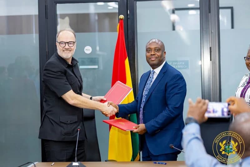 Dr. Mohammed Amin Adam, Ghana's Minister for Finance, shakes hands with a foreign official during a formal document signing ceremony. The two men are seen holding red folders in front of the Ghanaian flag within an office environment, likely at the Ministry of Finance.
