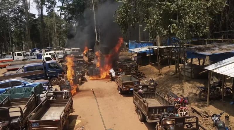 A clearing in a forest reserve is filled with numerous vehicles, including trucks and motor-tricycles, several of which are engulfed in flames. Thick black smoke and fire rise from the center of the assembly, which appears to be a site for illegal mining activities with makeshift shelters nearby.