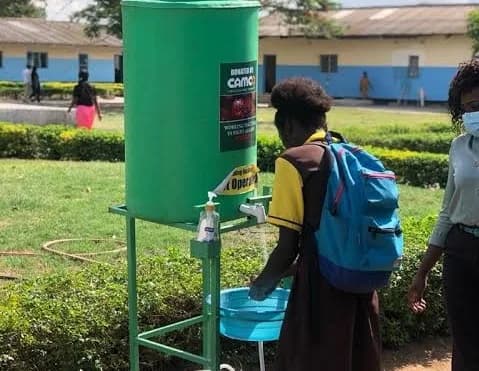 A student wearing a brown and yellow school uniform washes their hands at a green handwashing station, commonly known as a Veronica Bucket. A woman wearing a face mask stands to the right, and the station features a sticker indicating it was donated for the fight against COVID-19.