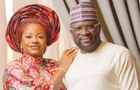 A photograph of Abdul-Wahab Hanan Aludiba, the former CEO of the National Food Buffer Stock Company (NAFCO), posing with a woman. Both are dressed in traditional attire; Hanan wears a white garment with a patterned kufi cap and glasses, while the woman wears a vibrant red patterned dress and a large matching headtie.