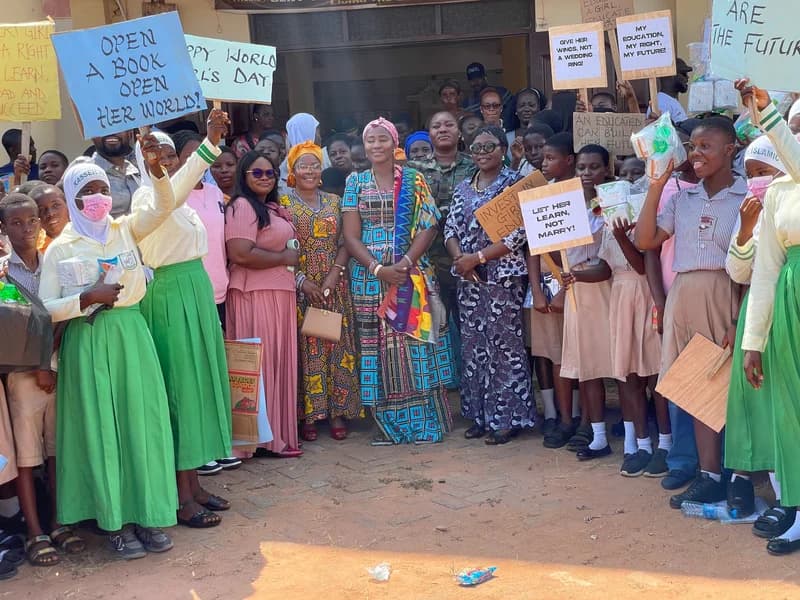 A group of Ghanaian school children and adults are gathered for an advocacy event focusing on girls' rights and education. Several participants are holding placards with messages such as 'Open a book, open her world!' and 'Let her learn, not marry!' to protest against child marriage.