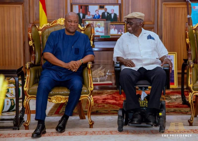 President John Dramani Mahama is pictured during a formal visit to former President John Agyekum Kufuor at his residence. The two leaders are seated together in a wood-paneled room, with Mahama in a blue traditional outfit and Kufuor in a white shirt and cap using a motorized wheelchair.