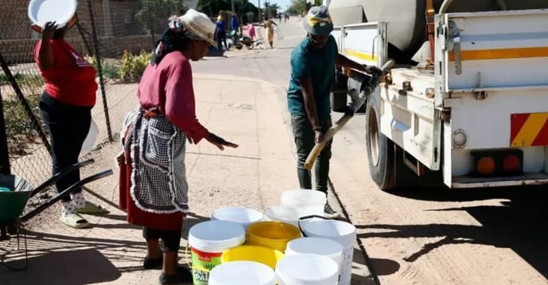 A man fills several white and yellow plastic buckets from a water tanker truck while residents wait nearby on a residential street. This scene illustrates the community's reliance on emergency water supplies following a health warning regarding contaminated tap water.