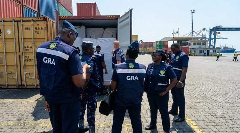 Officers from the Ghana Revenue Authority (GRA) Customs Division are pictured at a port or loading terminal standing before an open shipping container. The container is filled with large, white-wrapped bales, which likely contain the 560.4kg of seized cannabis mentioned in the report. The setting shows a busy industrial yard with numerous colorful shipping containers and port cranes in the background.