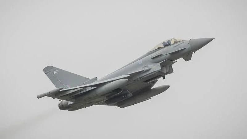 A grey Eurofighter Typhoon fighter jet is captured in mid-flight, climbing steeply against an overcast, grey sky. The aircraft is equipped with external fuel tanks and various munitions, illustrating a military deployment or patrol mission.