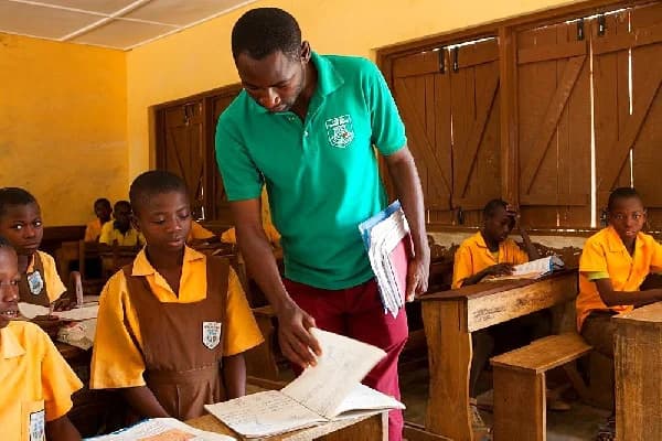 A male instructor wearing a green polo shirt is seen assisting a female primary school student with her work in a classroom setting. Several other students in yellow and brown school uniforms are visible in the background, seated at traditional wooden desks and engaged in reading.