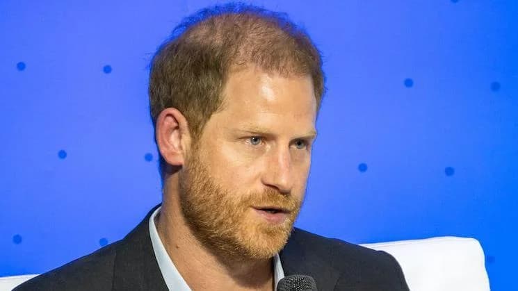 A close-up photograph of Prince Harry, Duke of Sussex, during a speaking engagement or interview. He is depicted with a ginger beard, wearing a dark blazer over a light-colored shirt against a solid blue backdrop.