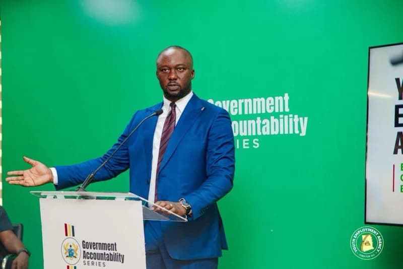 Kofi Baah Agyepong, the CEO of the Youth Employment Agency (YEA), is pictured delivering a presentation at a Government Accountability Series event. He stands behind a branded podium in a blue suit, gesturing as he speaks against a bright green background featuring event text.
