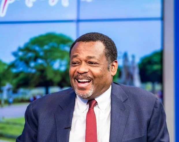 A portrait of Dr. Arthur C. Evans Jr. smiling during what appears to be a televised interview or presentation. He is wearing a navy blue suit, a white dress shirt, and a red tie, set against a studio background featuring a digital screen with an outdoor scene.