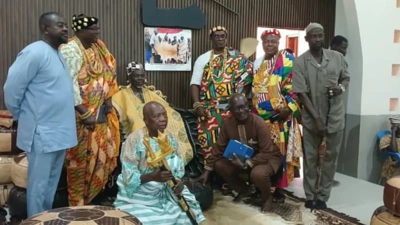 A group of Ghanaian traditional leaders and dignitaries are gathered for a formal occasion. A central figure sits holding a golden ceremonial staff while others stand or sit around him, most dressed in vibrant, multi-colored Kente cloth and traditional headgear.