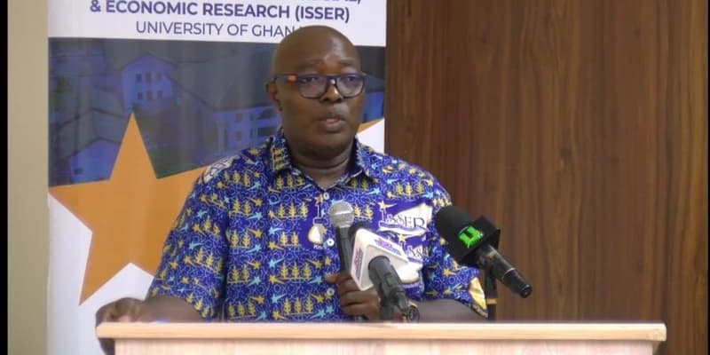 Professor Peter Quartey, Director of the Institute of Statistical, Social and Economic Research (ISSER), addresses a gathering at the University of Ghana. He is pictured speaking at a podium with several microphones, positioned in front of a banner representing the research institute.