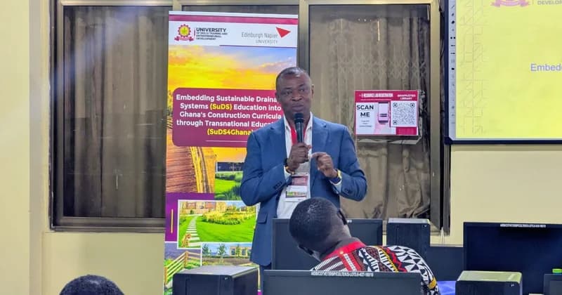 A professor at the University of Skills Training and Entrepreneurial Development (AAMUSTED) is seen delivering a presentation on sustainable drainage systems (SuDS). He is speaking into a microphone at an educational workshop focused on integrating these solutions into Ghana's construction curricula, standing before a banner and a digital screen.