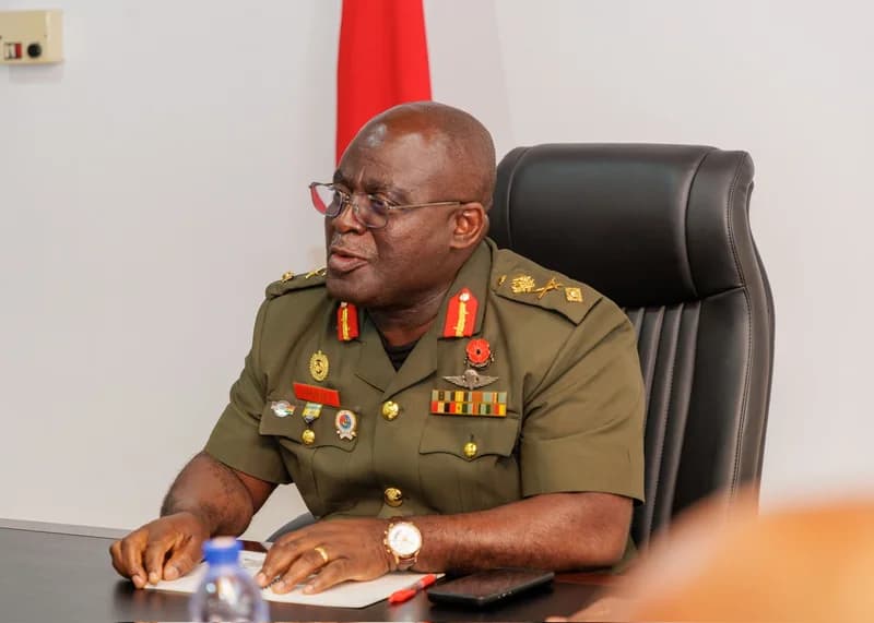 A high-ranking Ghanaian military official in an olive green service uniform is seated at a desk, appearing to be engaged in a formal discussion. He wears spectacles and has several service ribbons and a poppy pin displayed on his chest, seated in a black leather executive chair against a plain white background with a red flag.