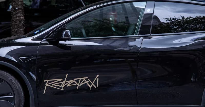 A close-up side view of a black Tesla vehicle featuring a prominent 'Robotaxi' decal in a stylized, graffiti-like beige font on the front door. The car's polished surface reflects a street environment, highlighting its role in Tesla's autonomous ride-hailing plans.
