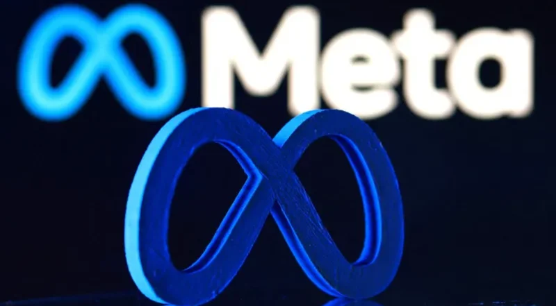The image features a physical 3D infinity logo representing Meta in the foreground, bathed in blue light. In the blurred background, the digital Meta logo and company name are visible on a screen against a dark backdrop.