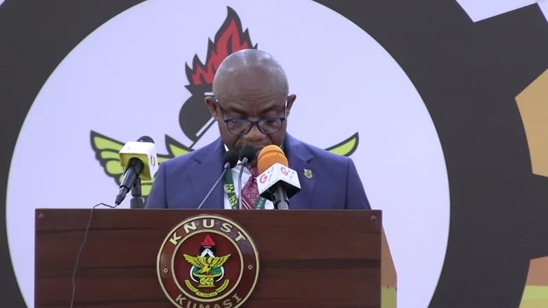 A man wearing glasses and a blue suit is pictured speaking at a wooden podium adorned with the KNUST Kumasi logo. Behind him is a large graphic display featuring the emblem of Kwame Nkrumah University of Science and Technology, during what appears to be an official university event.
