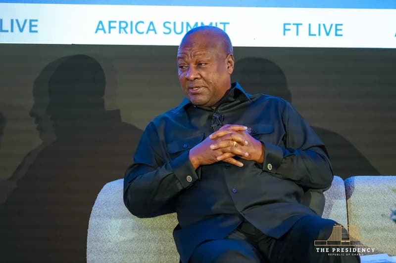 Ghanaian President John Dramani Mahama is captured speaking during a session at the FT Live Africa Summit. He is dressed in a black long-sleeved tunic, seated on a grey sofa with his hands clasped as he addresses the audience.